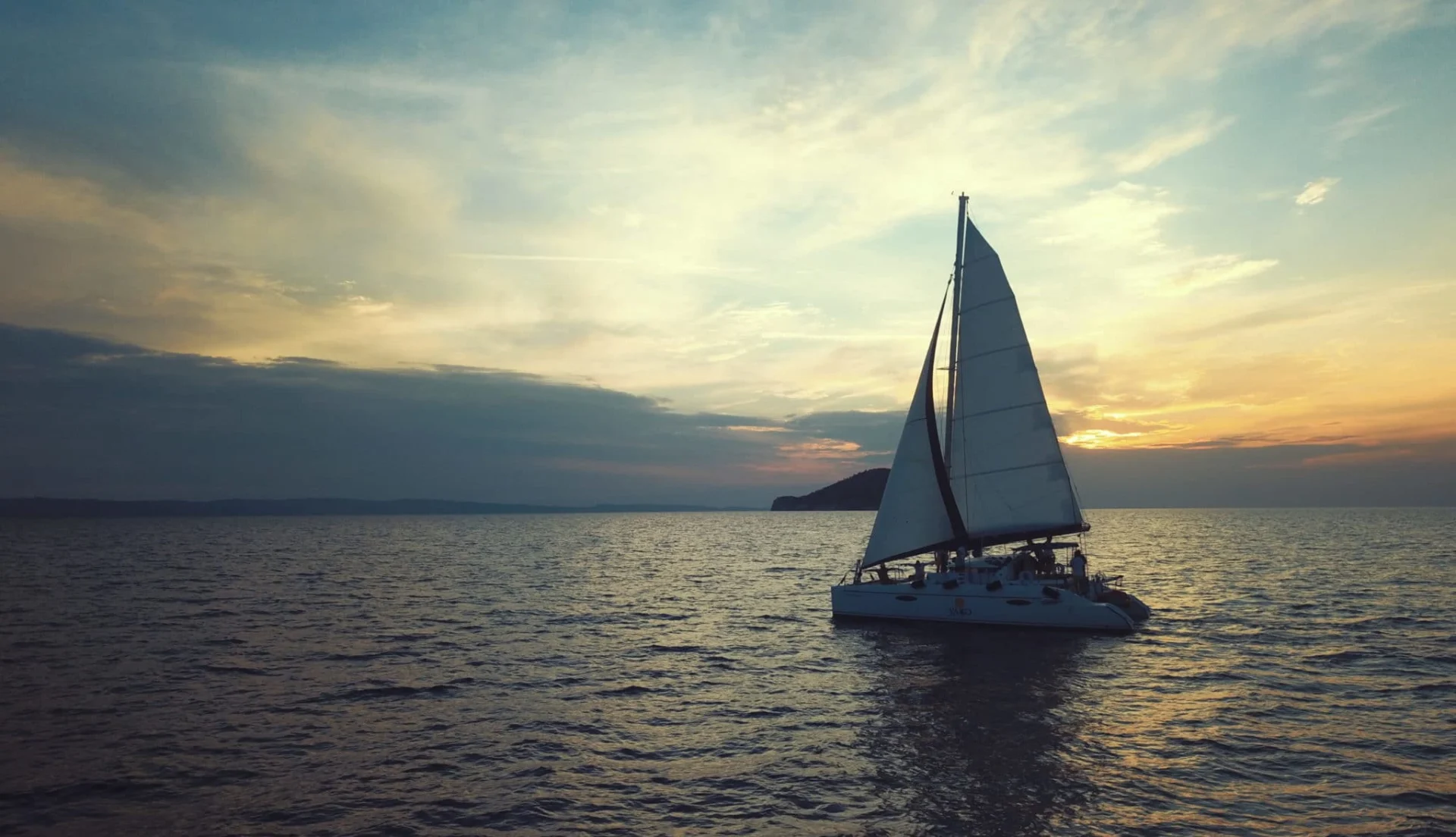 Catamaran under sail on a calm sea at sunset, with an island on the horizon