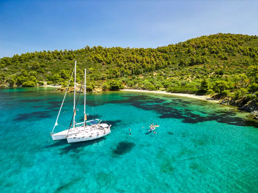 Two sailing yachts rafted together in a turquoise cove below pine-covered hills