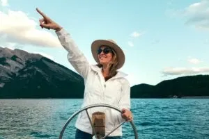 Smiling woman in a sun hat steering a boat and pointing ahead, mountains behind