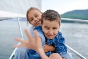 Two smiling children hugging and waving on the bow of a sailing boat