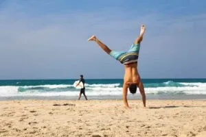 Man doing a handstand on a sandy beach while a surfer walks along the waves