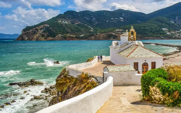 White church with a bell tower on a walled promontory above the sea, green hills behind