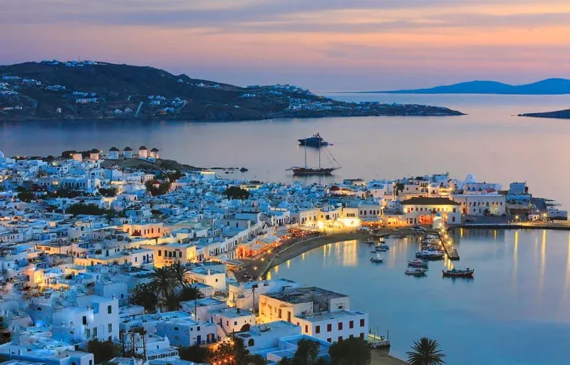 Mykonos town and its harbour lit up at dusk, with the hills beyond