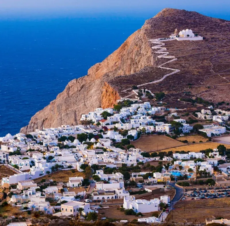White village below a steep hill with a zigzag path up to a church on Folegandros