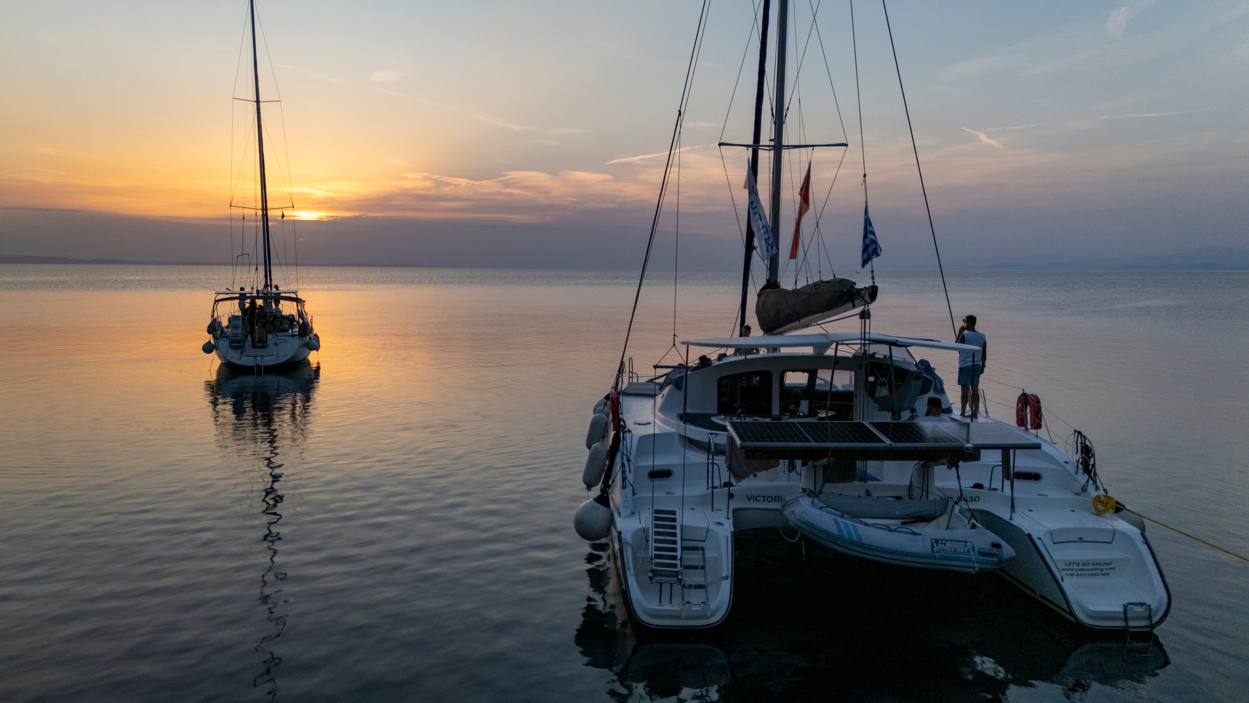 Catamaran and sailing yacht at anchor on calm water at sunset