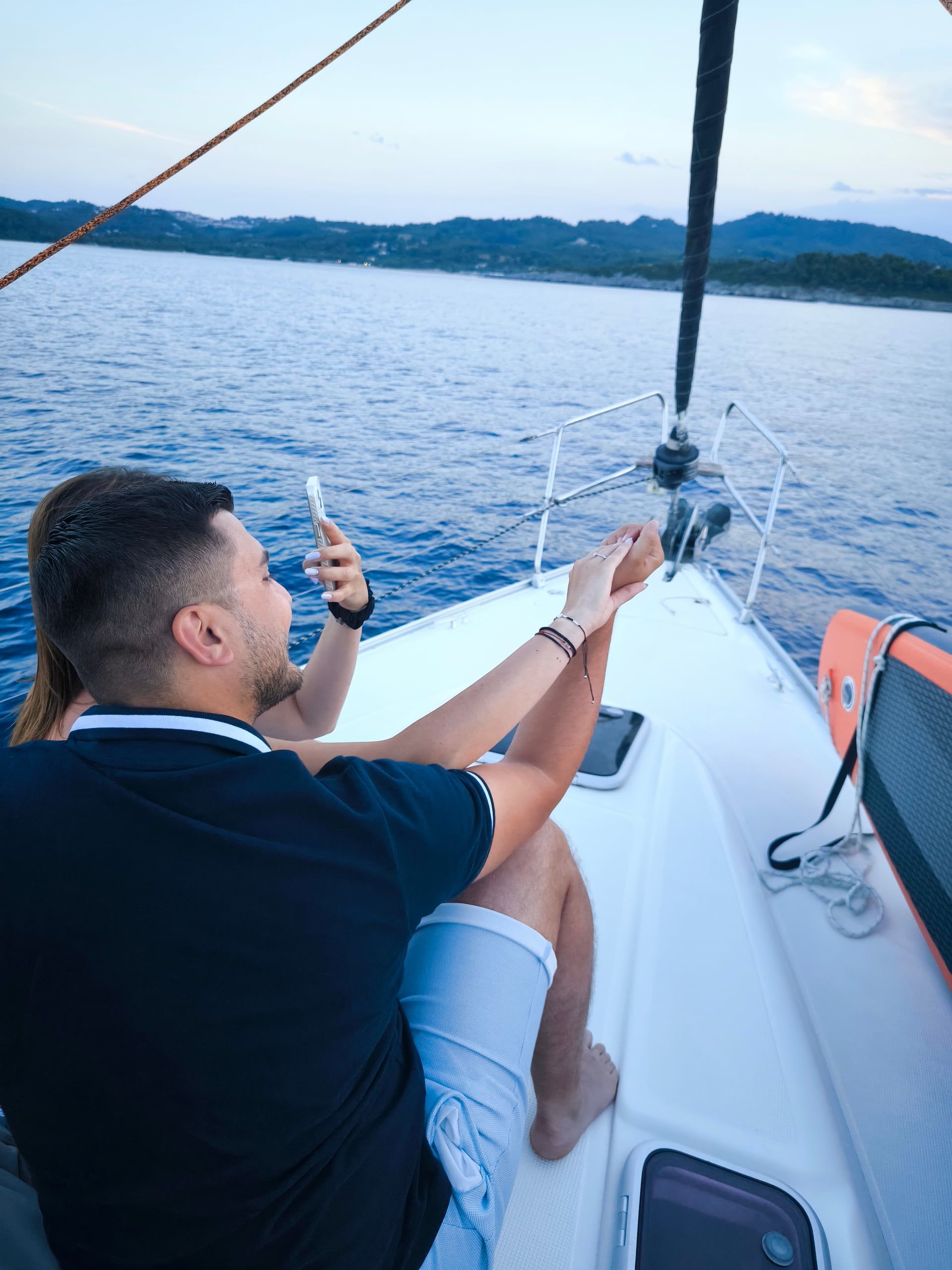 Couple on a sailing yacht’s bow photographing the coast at dusk