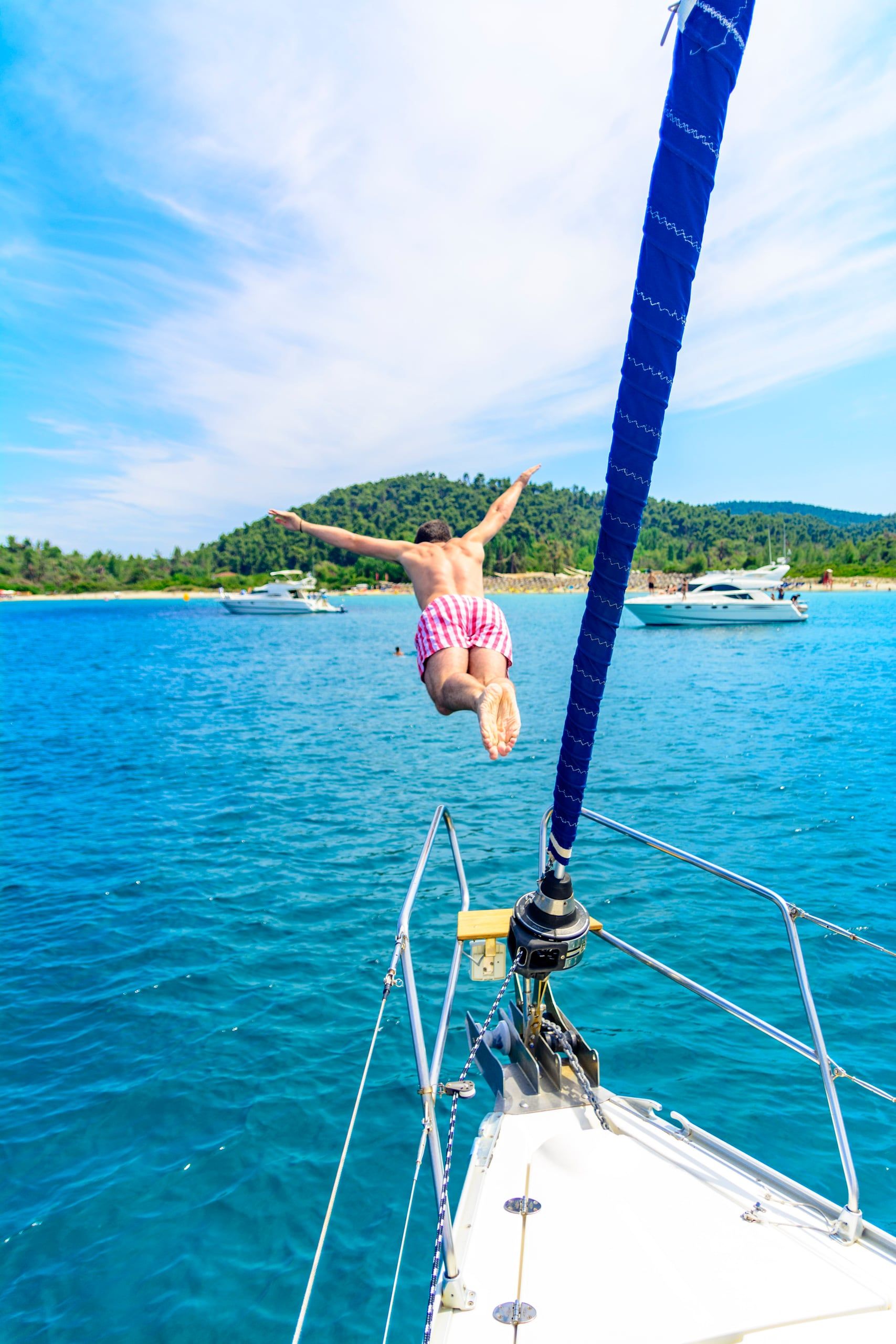 Man diving from a sailing yacht’s bow toward a wooded beach