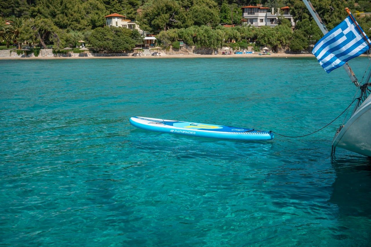 Paddleboard floating beside a sailing yacht with a Greek flag, villas on the shore