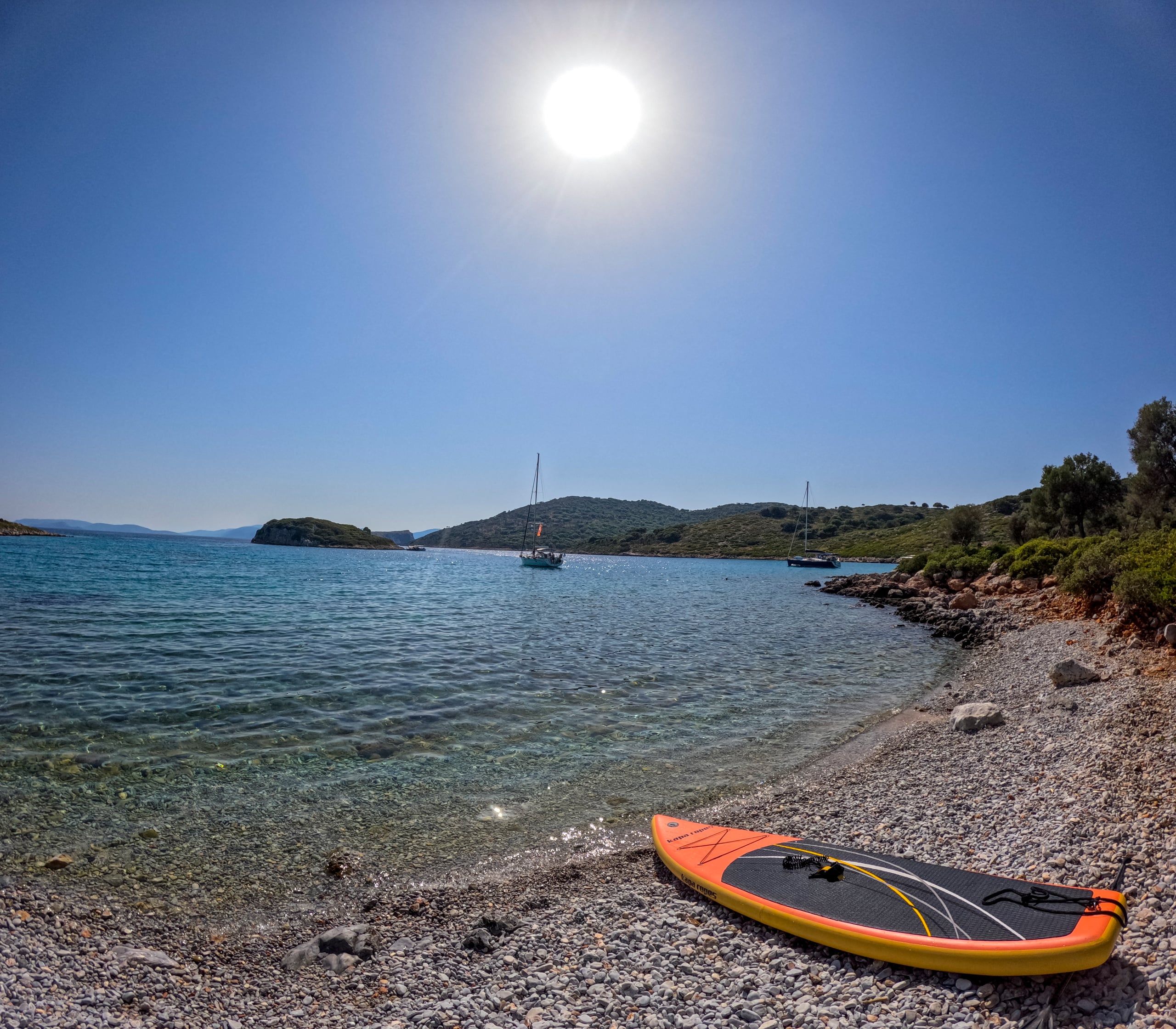 Orange paddleboard on a pebble beach, sailing yachts anchored in the bay