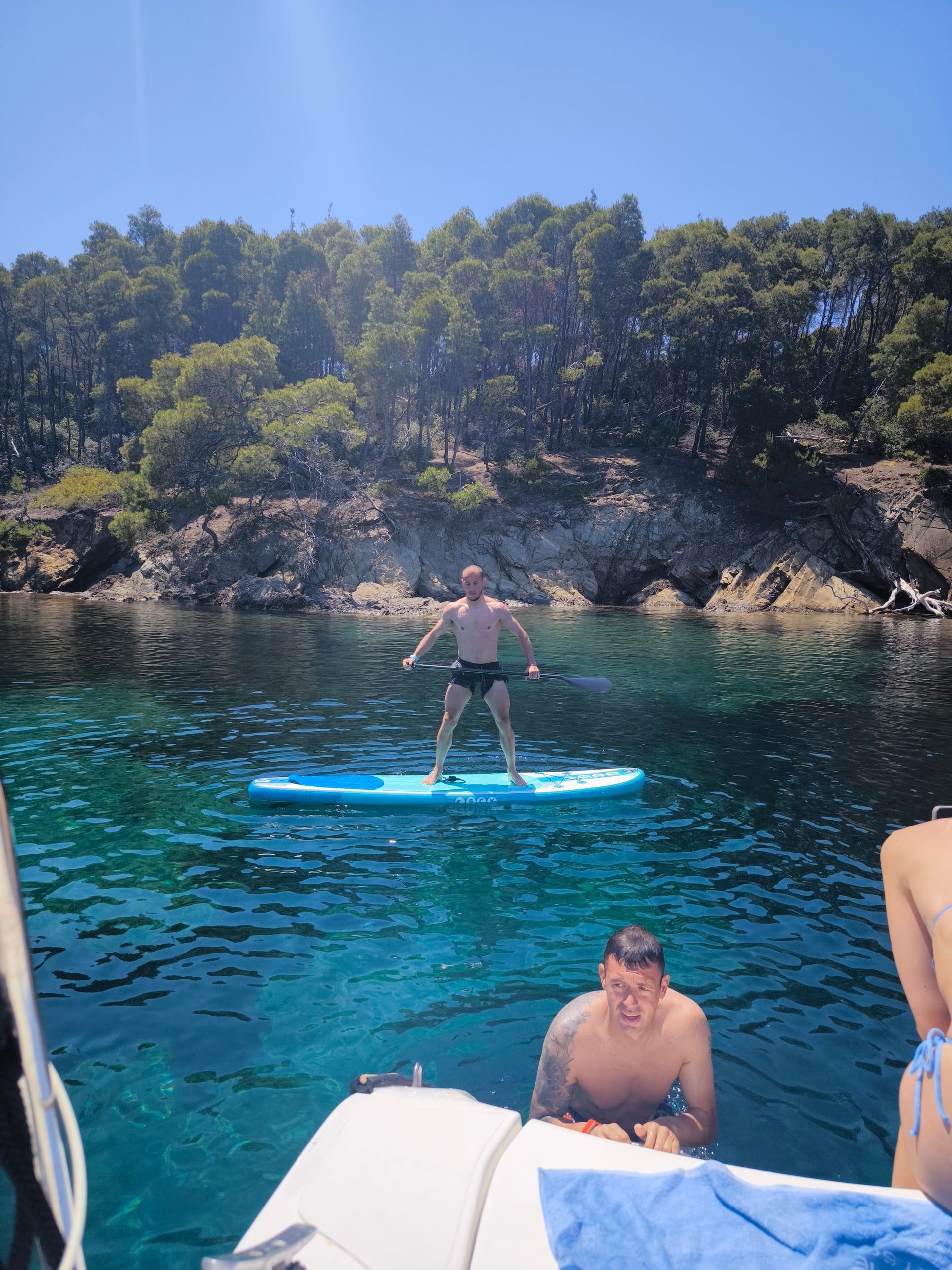 Guest standing on a paddleboard by a pine-covered rocky shore near Porto Carras
