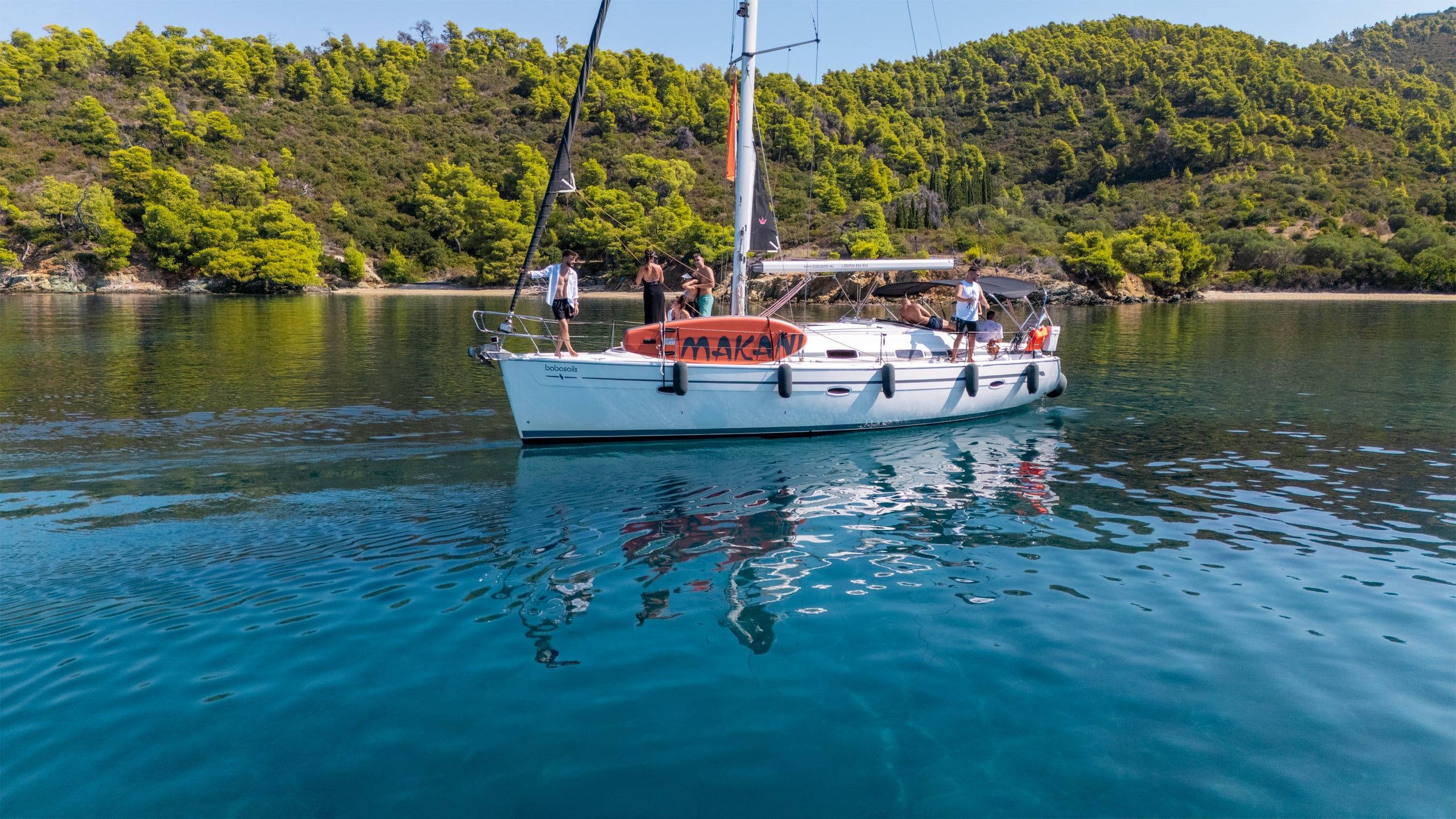 Guests on a sailing yacht at anchor in a calm bay below pine-covered hills near Porto Carras