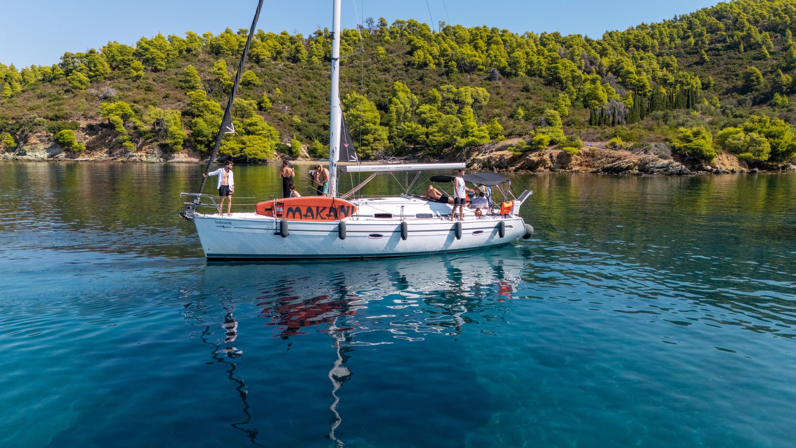 Guests on a sailing yacht with an orange paddleboard in a calm, pine-fringed bay