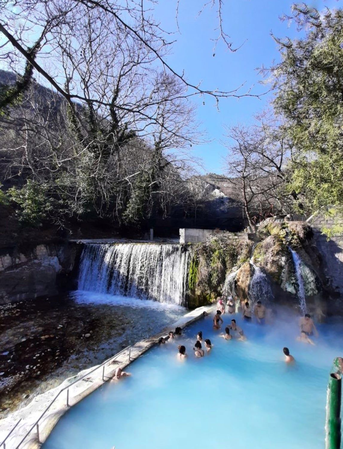 Steam rising over a bright blue thermal pool below a small waterfall at Pozar