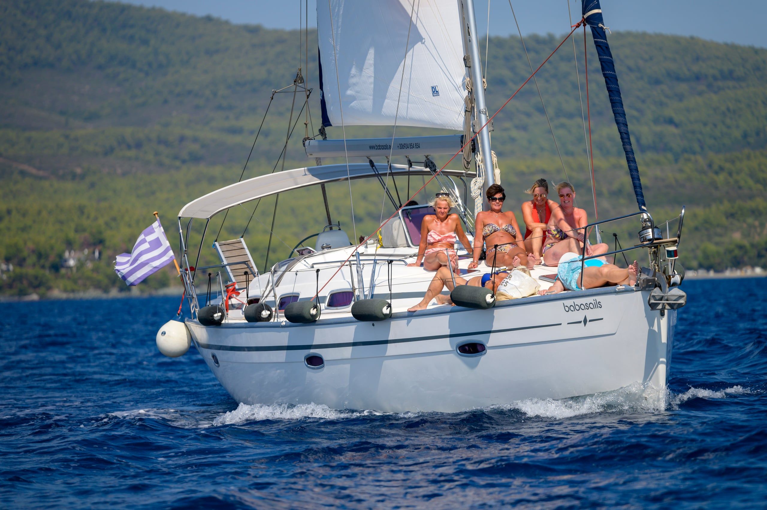 Sailing yacht under way with guests sunbathing on deck, wooded hills behind