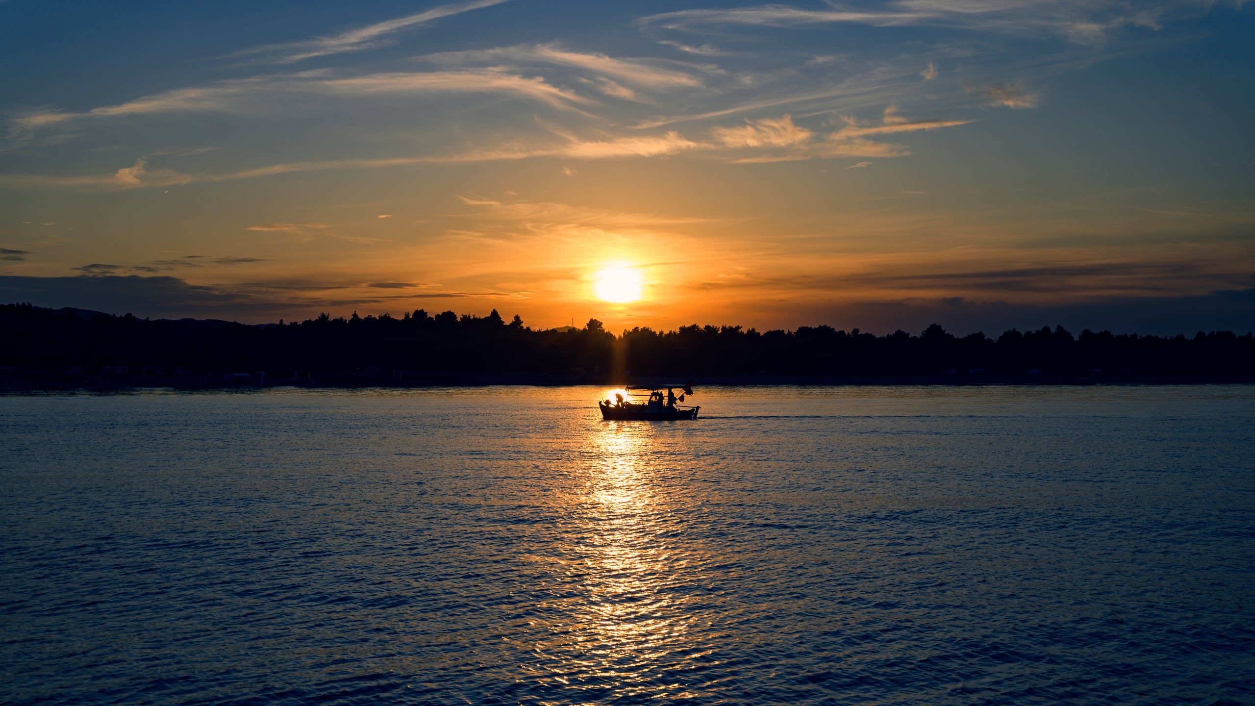 Small boat crossing a calm sea as the sun sets behind a wooded shore