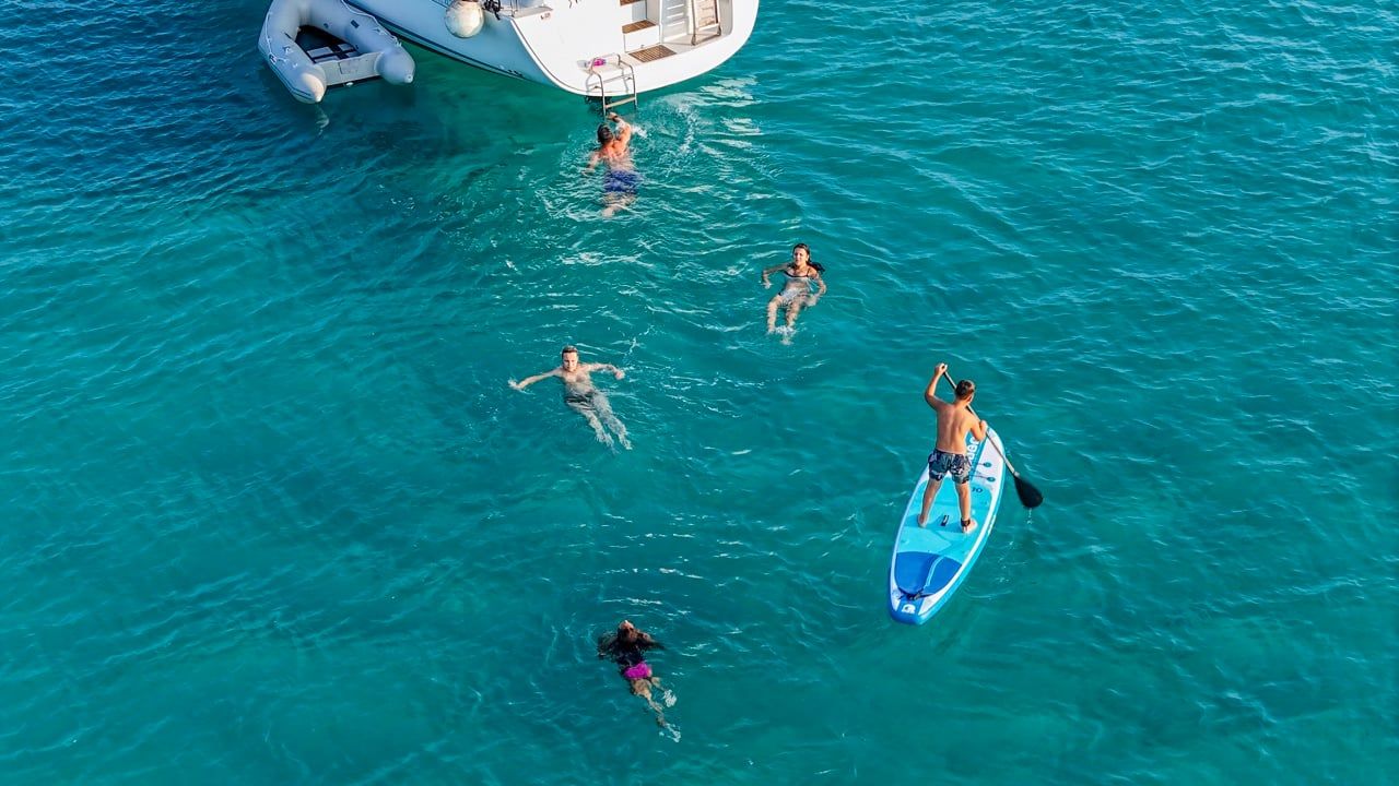 Swimmers and a paddleboarder in turquoise water beside a yacht, seen from above