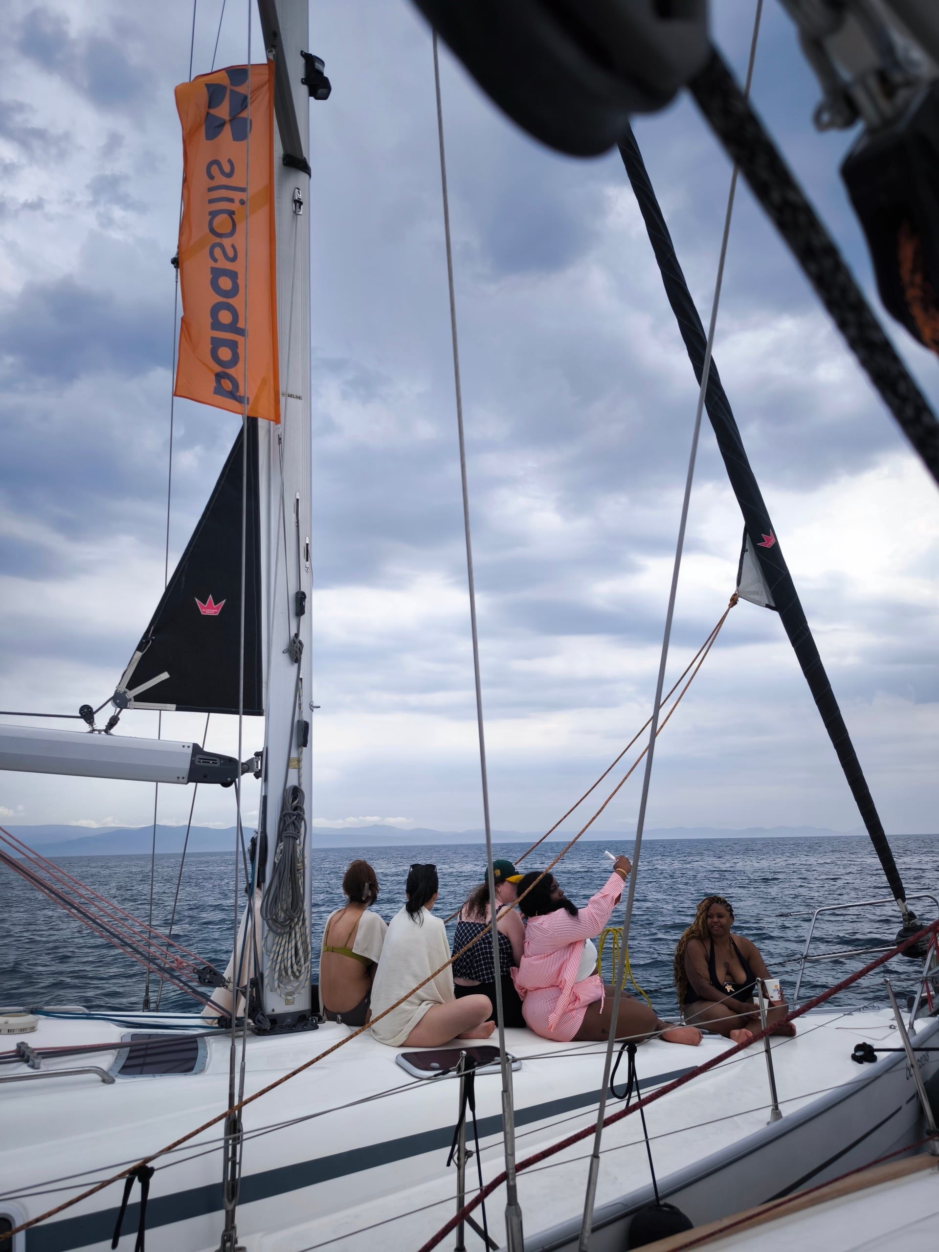 Women sitting on a sailing yacht’s bow under a cloudy sky, a flag on the mast
