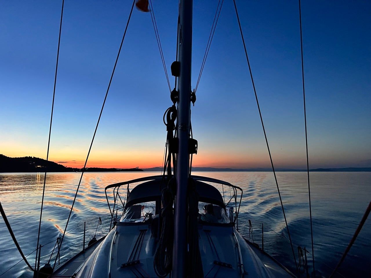 Calm sea at dusk seen from a sailing yacht’s bow, a glow on the horizon
