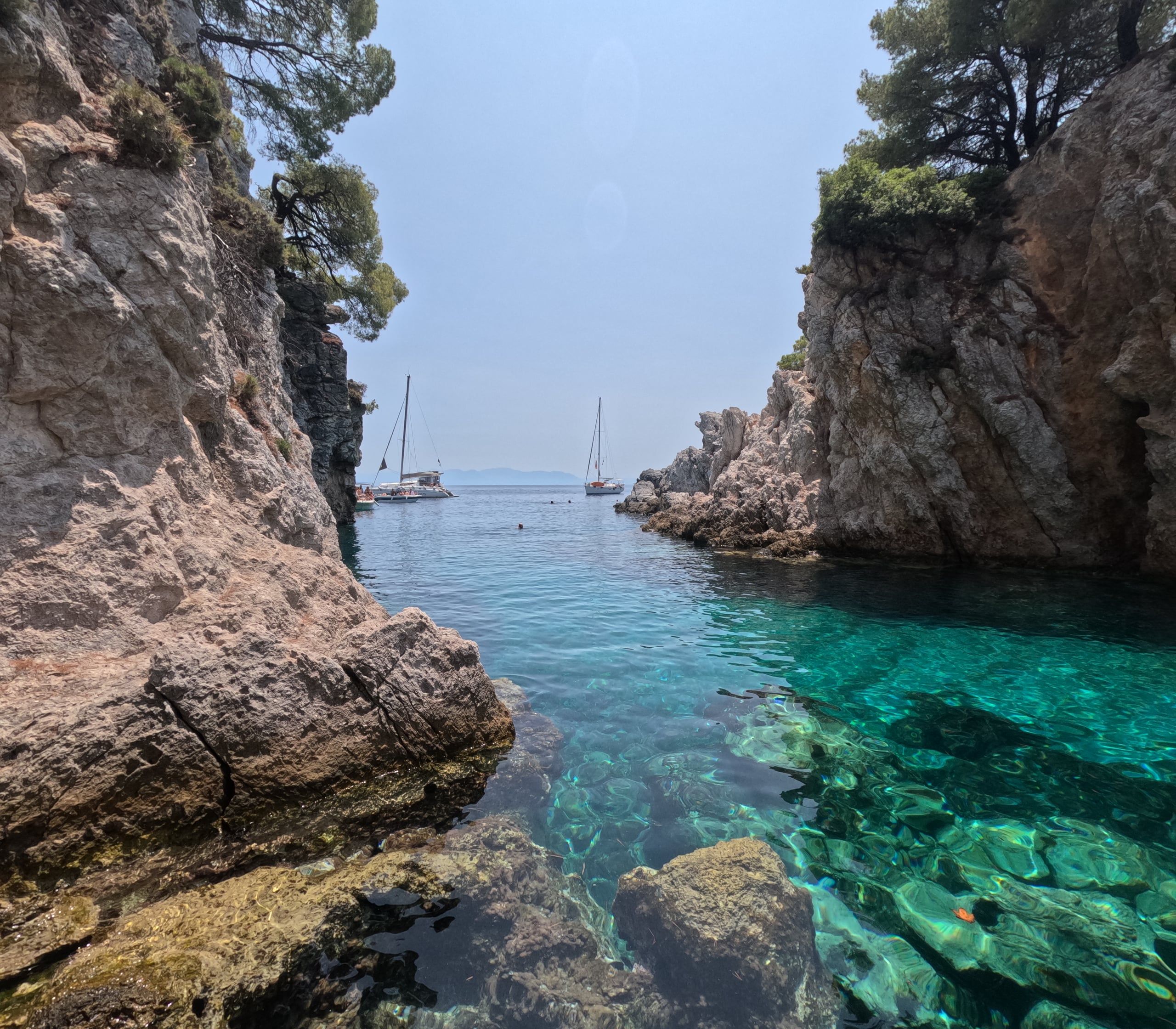 Two sailing yachts seen through a narrow inlet between rocky cliffs